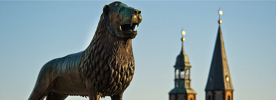 Bronzefigur des Löwes auf dem Braunschweiger Burgplatz vor dem Hintergrund eines blauen Himmel und zwei Kirchturmspitzen